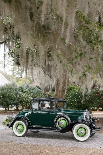 Vintage wedding car with green paint and whitewall tires parked on a gravel driveway beneath an oak tree with hanging moss