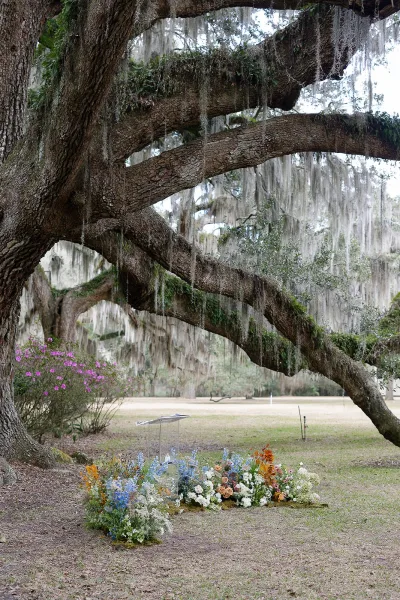 Outdoor ceremony setup beneath a moss-draped oak tree, with clear acrylic lectern and asymmetrical ground florals on a grassy lawn