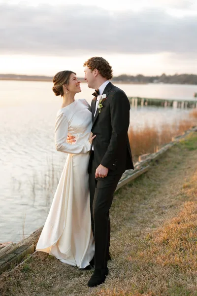 Couple portrait of bride and groom embracing on a waterfront dock, her long sleeve dress and pearl earrings under a cloudy sky