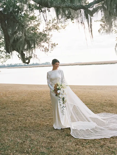 Bridal portrait of a bride holding bouquet of white orchids and greenery, wearing a long sleeve wedding dress and cathedral veil by an oak tree waterfront