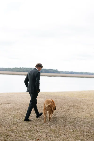 Groom with dog strolling by a lakeside field under an overcast sky, wearing a black suit and dress shoes beside a golden retriever