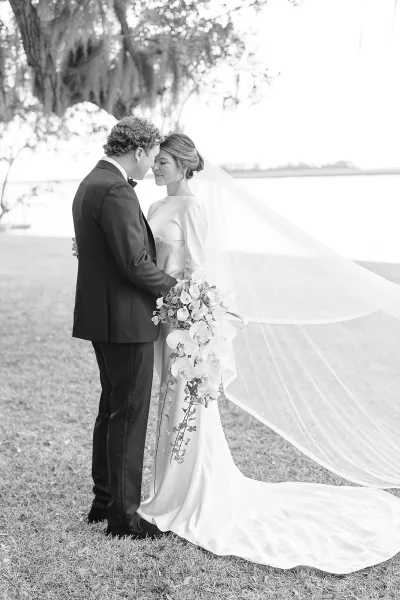 Couple portrait of wedding bride and groom touching foreheads as she holds an orchid bouquet by a waterfront lawn with trees and sky