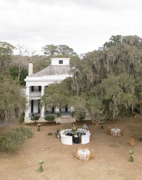 Outdoor reception setup with round tables and wooden chairs on an estate lawn, featuring a white bar by a historic porch under mossy trees