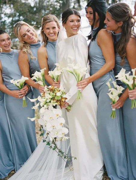 Bridal party portrait of a bride with bridesmaids in blue dresses, holding calla lilies and a cascading orchid bouquet outdoors by trees