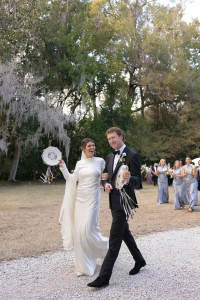 Wedding recessional as bride and groom walk the aisle, laughing under mossy trees; she lifts a ribbon tambourine as bridesmaids follow behind