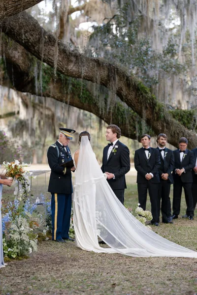 Wedding ceremony with bride and groom holding hands beneath an oak tree, long veil train flowing as a military officiant leads vows