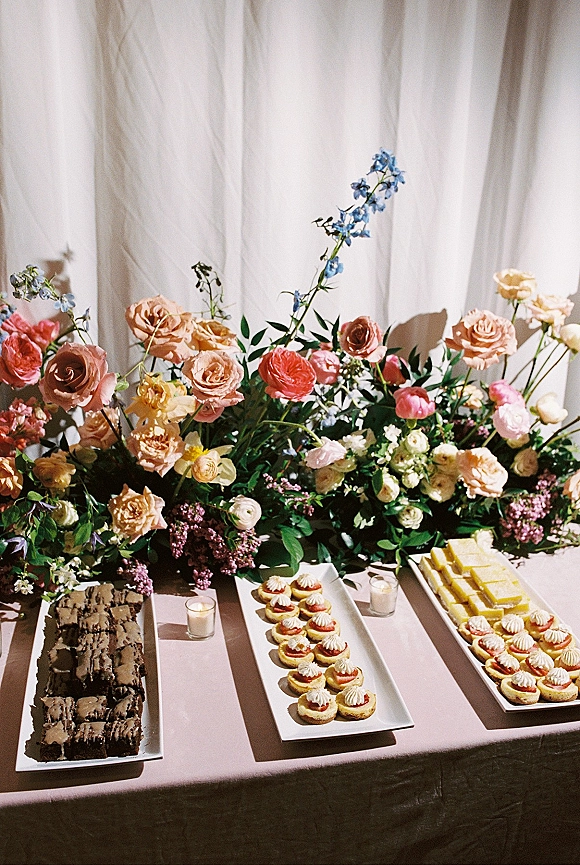 Wedding dessert table with mini tarts, brownie bars and petit fours on white platters, accented by roses, ranunculus and blue delphinium before white drapery backdrop