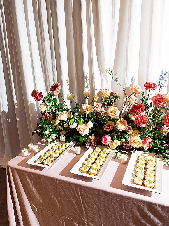 Dessert table with wedding dessert table decor of mini tarts on platters, roses and greenery with votive candles by sheer curtains in window light
