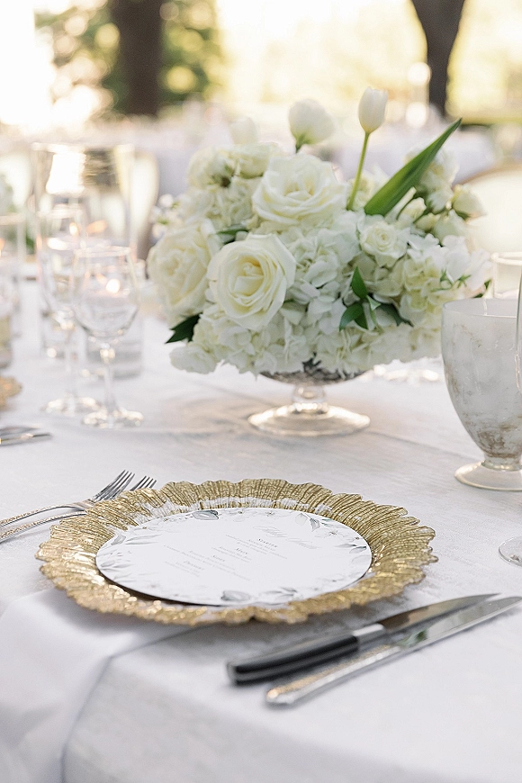 Reception tablescape with wedding table centerpiece of white roses, tulips and hydrangea in glass vase, gold chargers and candles outdoors