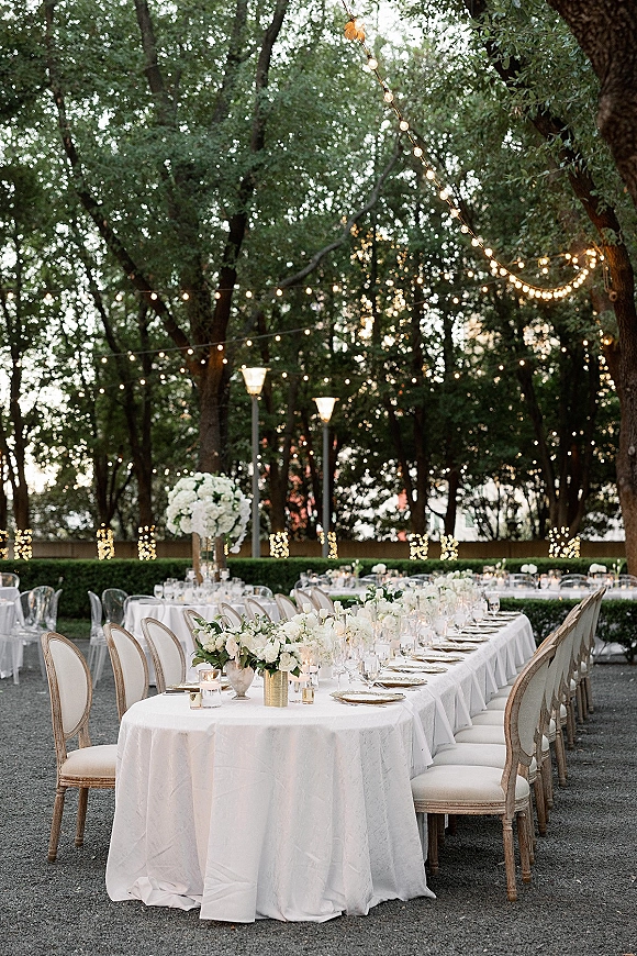 Reception tablescape for an outdoor wedding reception with a long white-linen banquet table, tall floral centerpieces, votive candles, and string lights under a tree canopy