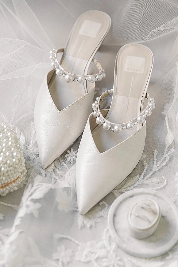 Wedding shoes with pearl ankle straps arranged on soft white textiles beside a ring box, wedding rings, and lace veil detail flat lay