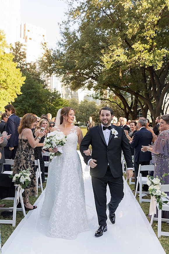 Wedding recessional as bride and groom walk the aisle, bride in lace dress and veil holding white bouquet in garden with guests cheering