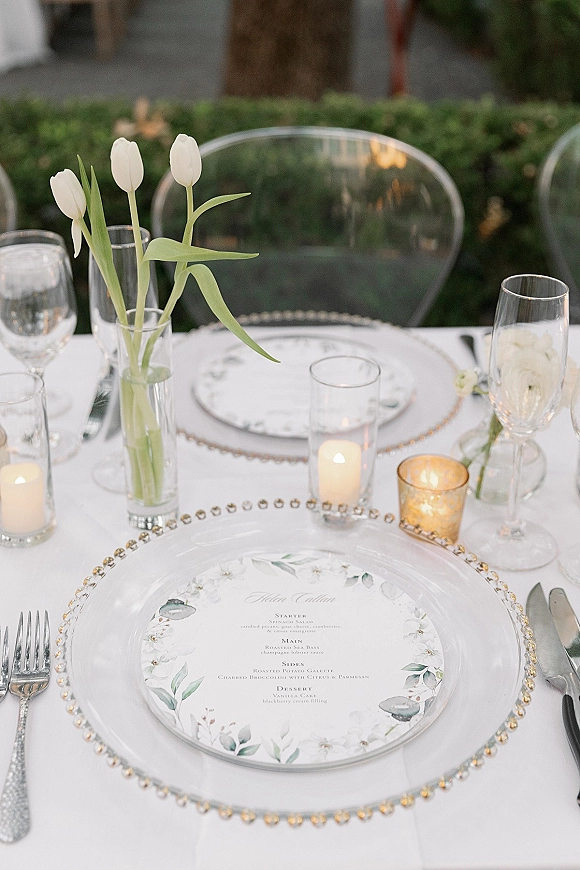 Reception tablescape with wedding place setting, beaded charger plate, menu card, pillar candles and white tulips on an outdoor patio by a hedge