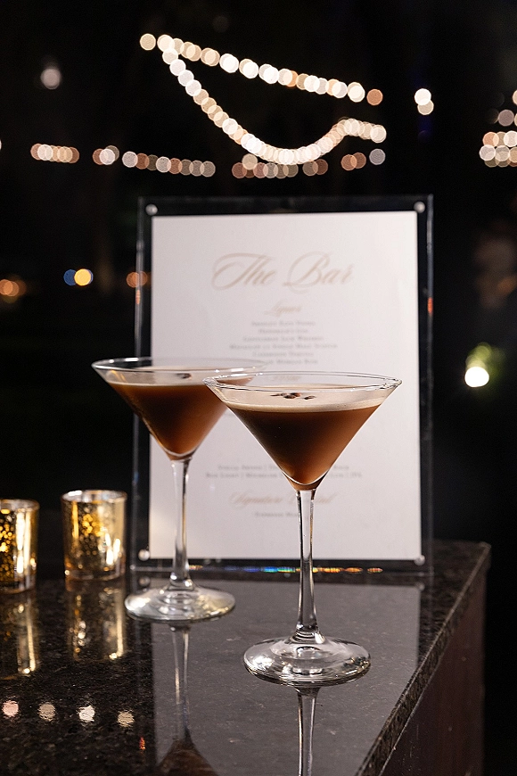 Wedding bar menu with signature cocktail menu in a clear acrylic holder beside martini glasses and votive candles under string lights at night