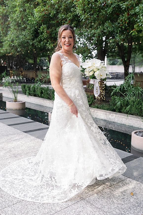 Bridal portrait of a laughing bride holding a white rose and orchid bouquet, wearing a lace gown with long train in a garden courtyard