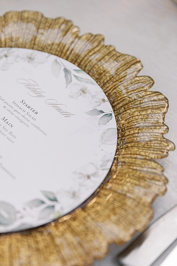 Wedding menu card with calligraphy wedding menu design, set on a scalloped gold charger plate atop crisp white table linen with floral illustration