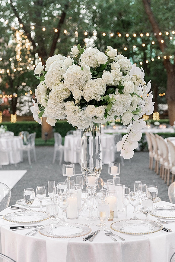 Reception tablescape with a tall floral centerpiece of white hydrangeas, roses and orchids in a glass vase at an outdoor garden under string lights