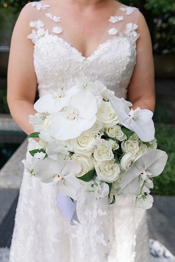 Bridal bouquet with a white orchid bouquet of orchids and roses, greenery and ribbon, held against a lace illusion-neckline dress on stone steps