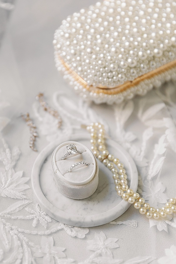 Wedding rings with an engagement ring close up on a marble dish beside a pearl necklace and lace fabric on a soft neutral surface