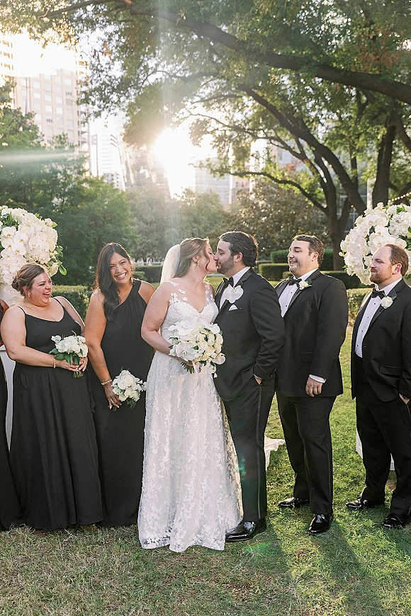 Wedding party portrait of bride and groom kissing as bridesmaids in black dresses and groomsmen in tuxedos stand on a sunlit city park lawn with trees