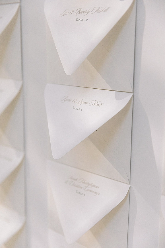 Wedding escort cards in a wedding seating cards display, white folded paper with calligraphy arranged on a wall with soft shadows