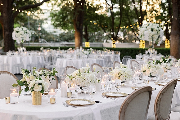 Reception tablescape with white floral centerpieces and candlelit place settings, gold charger plates under string lights in a garden at dusk