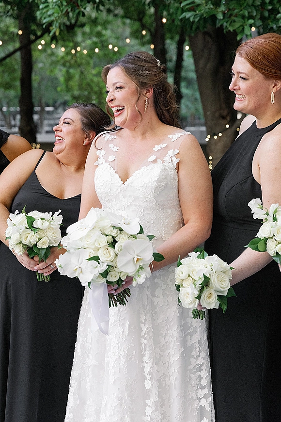 Bridesmaid group photo with bride and bridesmaids laughing in black dresses, holding white rose bouquets under garden string lights