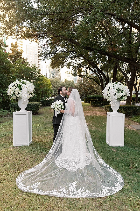 Wedding kiss portrait of bride and groom kissing as her cathedral veil and lace train flow, holding a bouquet on a park lawn with trees and floral pedestals