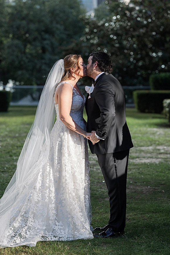 Wedding kiss portrait of bride and groom kissing, holding hands; lace dress and veil with groom tuxedo on lawn by trees and building