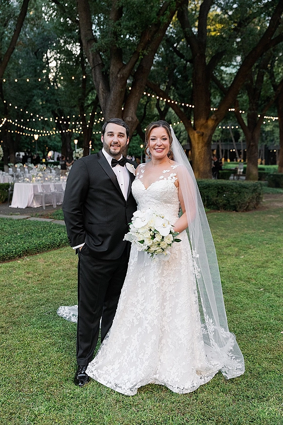 Couple portrait of bride and groom portrait smiling under large trees, her veil and white bouquet beside his black tuxedo with string lights behind