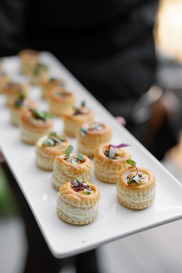 Wedding appetizers arranged as passed hors d'oeuvres, mini puff pastry bites topped with microgreens on a white platter at a blurred reception