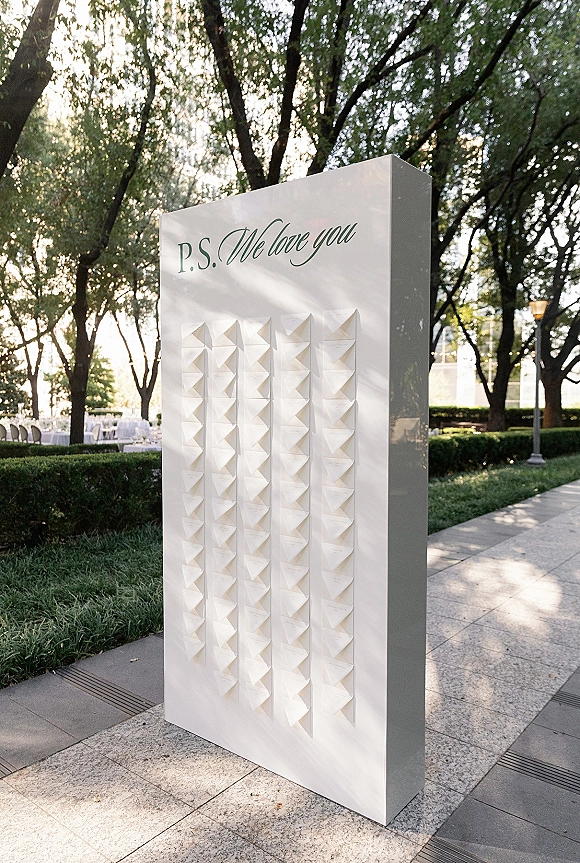 Wedding escort card display with calligraphy escort cards on a white board in an outdoor garden setting with string lights and hedges