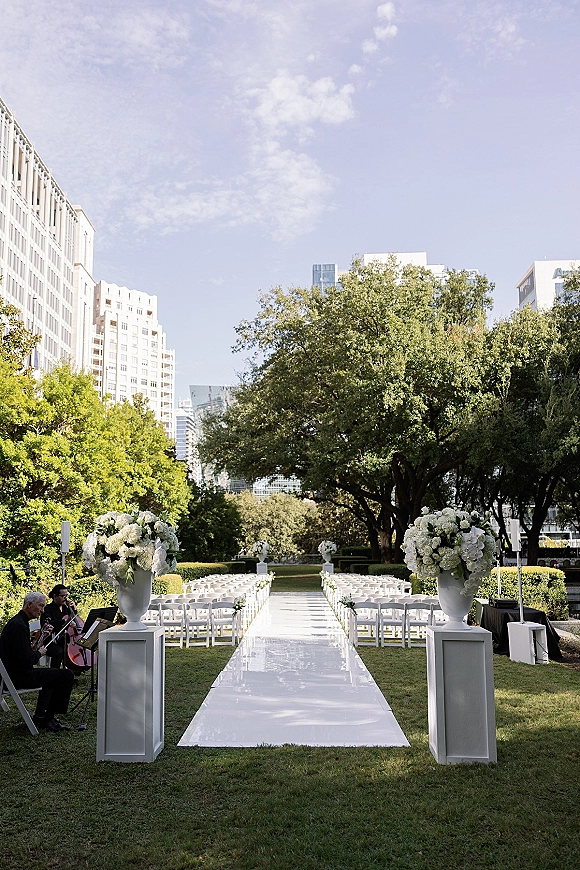 Ceremony aisle decor with a white aisle runner flanked by white folding chairs and floral urn pedestals on a city park lawn under blue sky