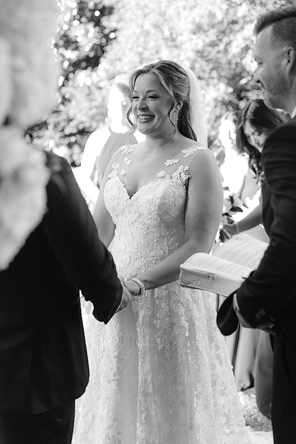 Wedding vows as bride and groom hold hands, bride smiling at groom while officiant reads from a book amid outdoor greenery guests