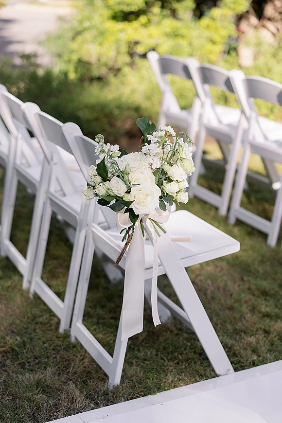 Ceremony aisle decor with wedding chair flowers on white folding chairs, white rose arrangements tied with a ribbon bow on a sunny lawn