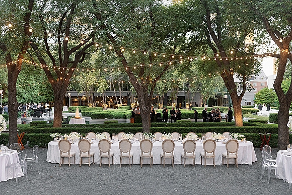 Reception tablescape with an outdoor reception table set on a long white-clothed banquet, white florals, greenery, candles, and string lights in a garden courtyard