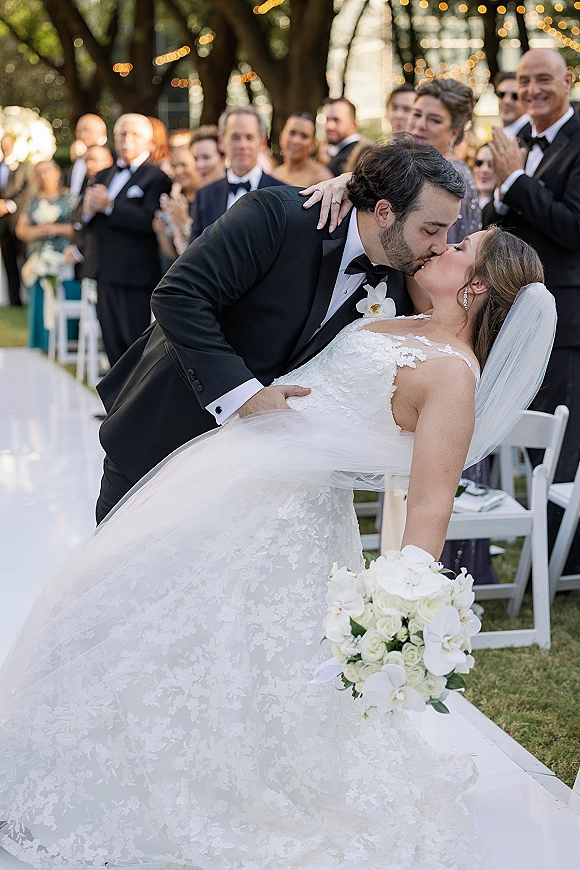 Wedding kiss portrait of bride and groom in a dip kiss wedding photo, cathedral veil and orchid bouquet as guests clap in garden aisle