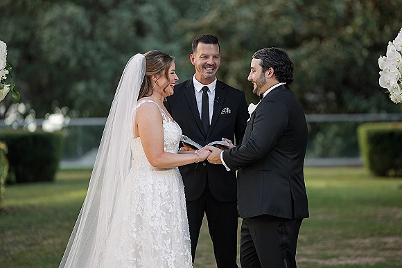 Wedding vows shared as bride and groom hold hands, her lace dress and long veil beside tuxedoed groom on a garden lawn with trees