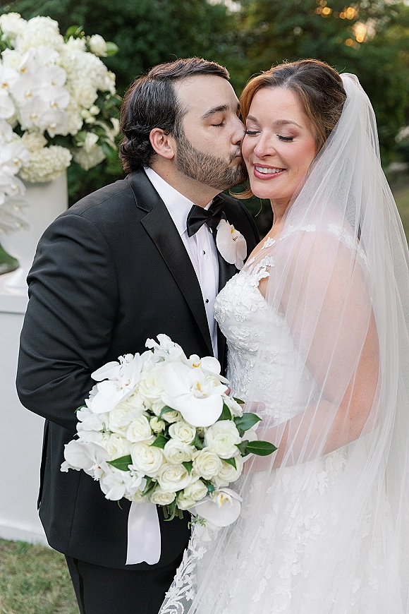 Wedding couple portrait with groom kissing bride’s cheek as she smiles in veil, holding white rose and orchid bouquet in garden greenery