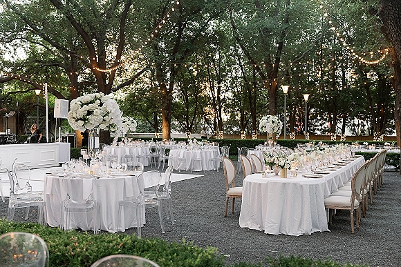 Outdoor reception decor with string light wedding reception glow over white floral centerpieces, candles, and place settings in a garden courtyard