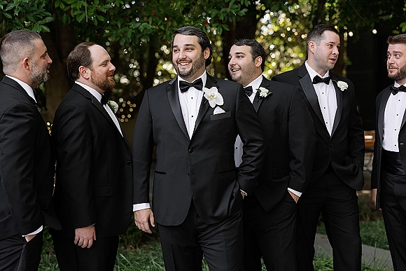 Groomsmen portrait of the groom and groomsmen in black tuxedos with white orchid boutonnieres on an outdoor garden walkway under string lights