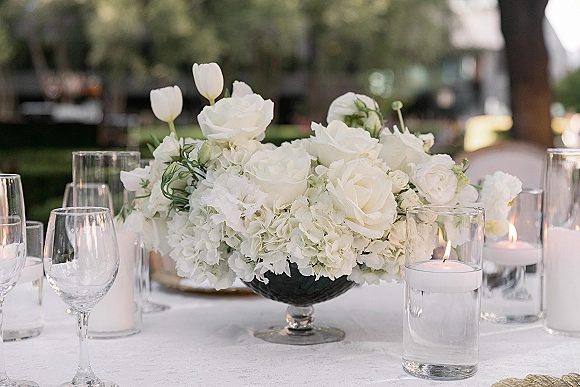 Reception centerpiece with a white floral centerpiece of roses, tulips, and hydrangea in a compote bowl, with floating candles on a garden table