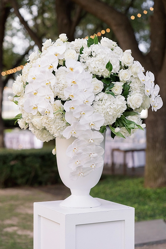 Wedding floral arrangement in a white urn centerpiece with cascading orchids, roses and hydrangeas on a garden pedestal under string lights