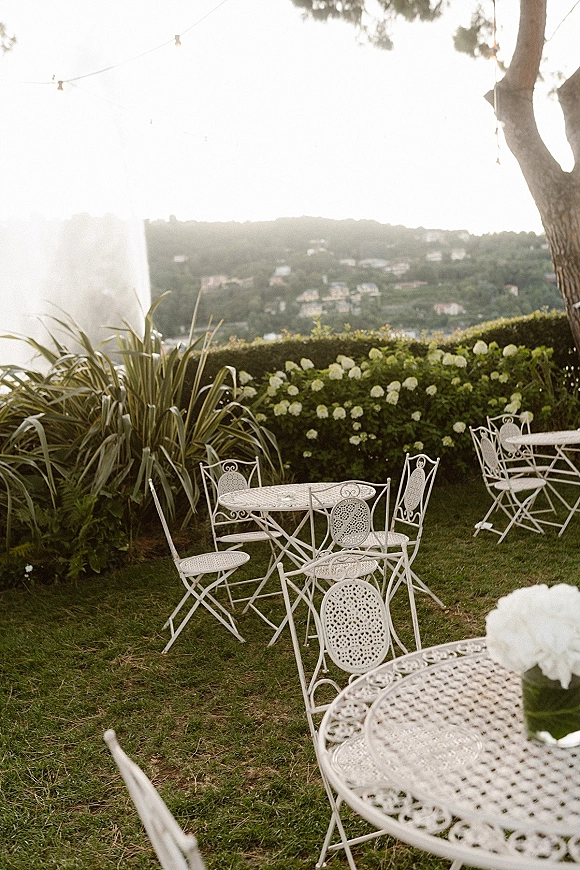 Outdoor reception seating with white wrought iron bistro tables and hydrangea centerpieces under string lights on a garden lawn hillside view