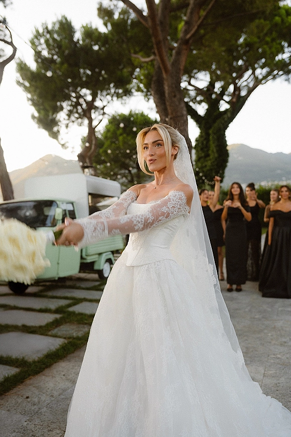 Bouquet toss as the bride tossing bouquet in a lace off-the-shoulder gown and long veil, bridesmaids in black watch in a garden with mountains