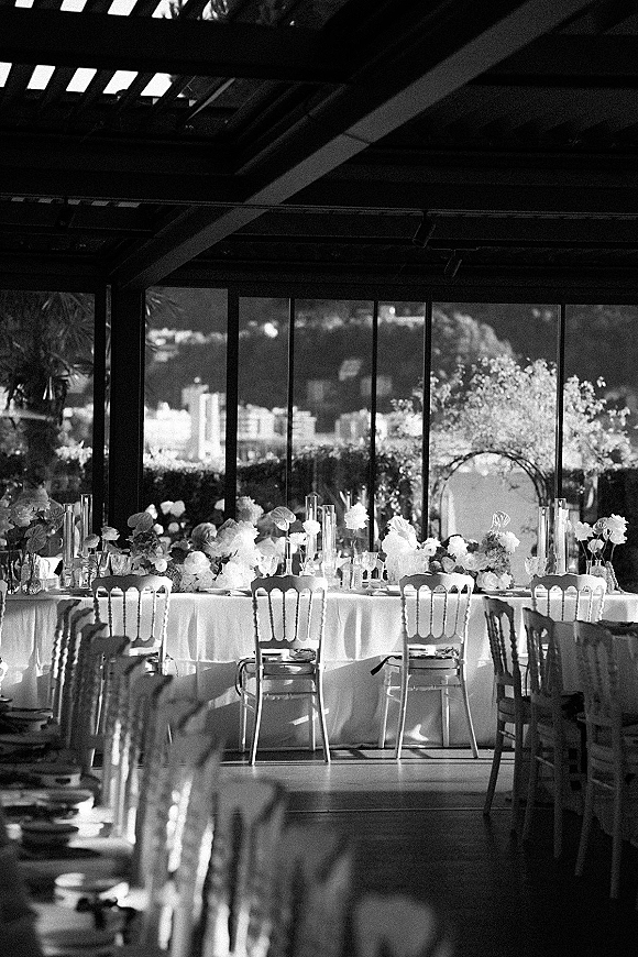 Reception tablescape with head table decor on a long white-clothed banquet table, white floral centerpieces and candles by glass wall windows