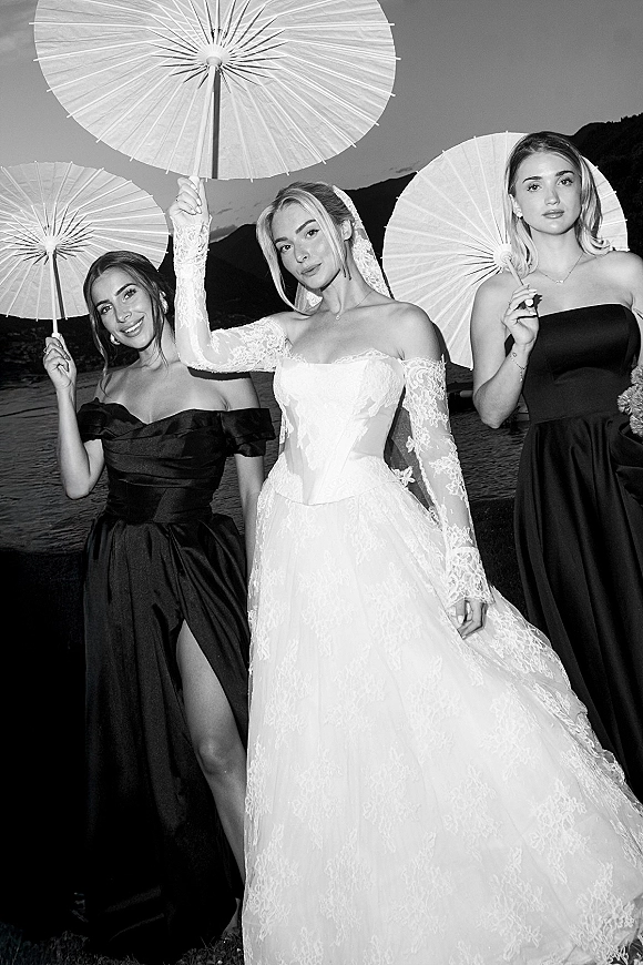 Bride with bridesmaids holding paper parasols, bride in lace-sleeve gown and veil with bouquet by a lake and mountain silhouette backdrop