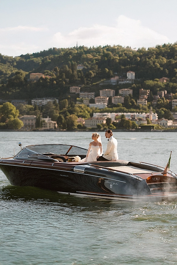 Couple portrait of bride and groom on boat, her veil and wedding dress flowing beside his tuxedo in a wooden speedboat on a lake