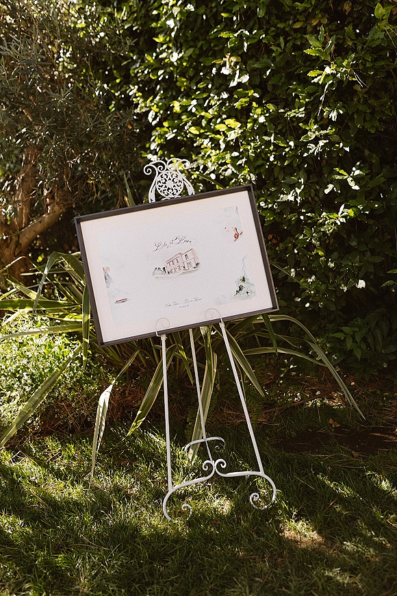 Wedding welcome sign in a framed display on a white metal easel, featuring illustrated artwork, set against garden greenery in dappled sunlight