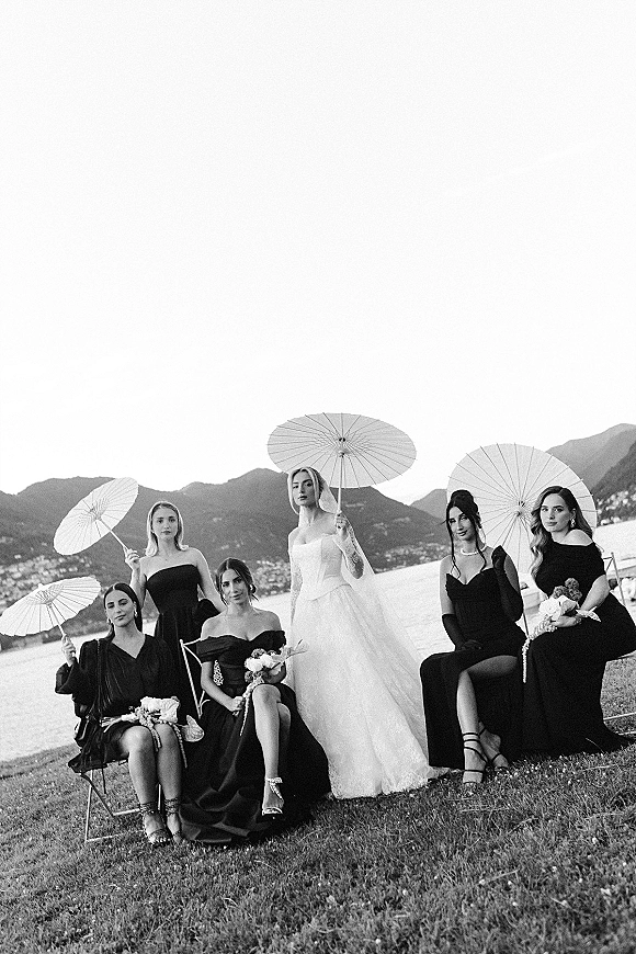 Bridal party portrait of bride with bridesmaids holding paper parasols and bouquets, seated on chairs by a mountain lake shoreline